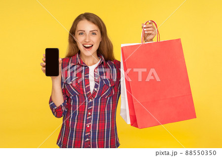Amazed surprised ginger girl in casual style checkered shirt holding shopping bags and mobile phone, advertising online store, looking shocked. indoor studio shot isolated on yellow background Amazed surprised ginger girl in casual style checkered shirt holding shopping bags and mobile phone, advertising online store, looking shocked. indoor studio shot isolated on yellow background 88550350