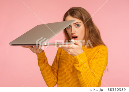 Portrait of astonished ginger girl in sweater holding half-opened laptop and looking at camera with surprise amazement, shocked by forbidden bad content. indoor studio shot isolated on pink background 88550389