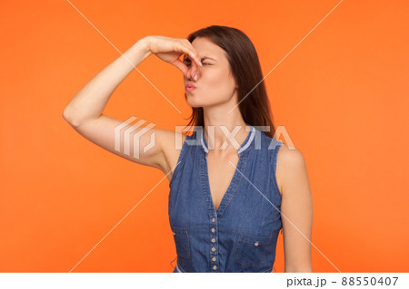 Stink, awful odor. Brunette woman pinching nose, stop breathing bad odor, disgusted by smell of farting, her grimace expressing repulsion, gross. indoor studio shot isolated on orange background Stink, awful odor. Brunette woman pinching nose, stop breathing bad odor, disgusted by smell of farting, her grimace expressing repulsion, gross. indoor studio shot isolated on orange background 88550407