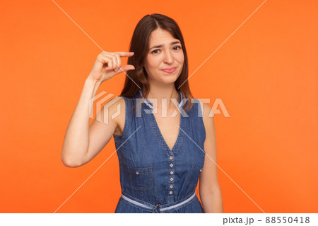 Small scale. Positive brunette woman in denim dress showing a little bit gesture and looking with disappointment, measuring centimeter, inch size. indoor studio shot isolated on orange background 88550418