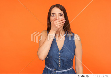 Scared brunette woman in denim dress covering mouth with hand and looking frightened at camera, afraid to talk, freedom of speech censorship concept. indoor studio shot isolated on orange background 88550520
