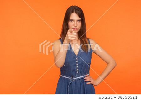 Hey you. Strict displeased brunette woman in denim dress pointing finger to camera and looking with suspicious angry expression, making accusing gesture. studio shot isolated on orange background 88550521
