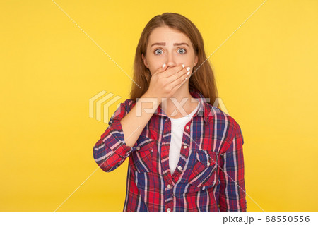 I won't tell anyone. Portrait of scared ginger girl in checkered shirt covering mouth with hand and looking frightened at camera, intimidated female afraid to talk. studio shot, yellow background I won't tell anyone. Portrait of scared ginger girl in checkered shirt covering mouth with hand and looking frightened at camera, intimidated female afraid to talk. studio shot, yellow background 88550556