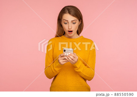 Portrait of surprised cute ginger girl texting on cellphone and expressing amazement, reading shocking message using smartphone, mobile application. indoor studio shot isolated on pink background 88550590