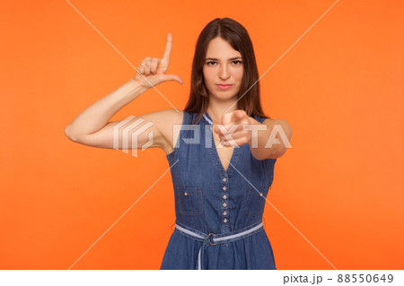 Bossy woman in denim dress showing L sign and pointing to camera, gesturing you are lame loser, mocking insulting for defeat, looking with disrespect. indoor studio shot isolated on orange background 88550649