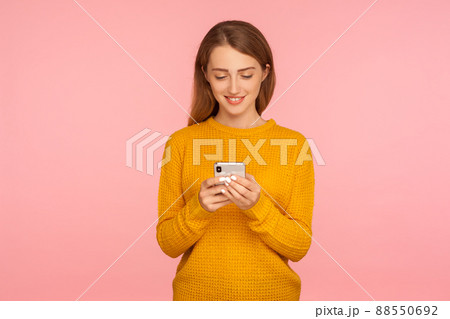 Portrait of positive cute ginger girl typing message on cellphone and smiling, scrolling social network or shopping using smartphone, mobile application. indoor studio shot isolated on pink background Portrait of positive cute ginger girl typing message on cellphone and smiling, scrolling social network or shopping using smartphone, mobile application. indoor studio shot isolated on pink background 88550692