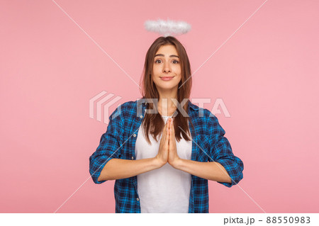Portrait of angelic kind pretty girl with nimbus over head holding hands in prayer gesture and looking at camera with saint trustworthy expression. indoor studio shot isolated on pink background 88550983