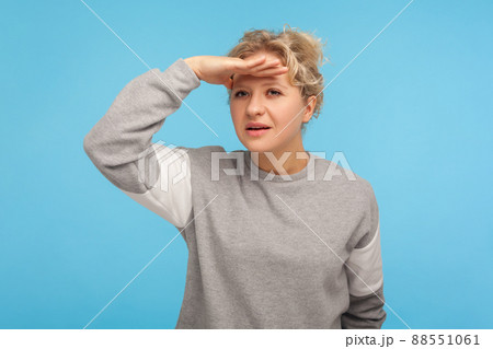 Distant view gesture. Woman with curly hair in sweatshirt attentively looking far away, keeping hand above eyes watching forward future, perspectives. indoor studio shot isolated on blue background 88551061
