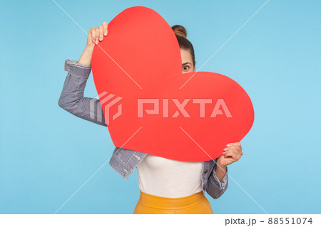 Portrait of funny optimistic woman peeking out of big red paper heart in her hands, looking with curious and amazed big eye, surprise on Valentines day. indoor studio shot isolated on blue background Portrait of funny optimistic woman peeking out of big red paper heart in her hands, looking with curious and amazed big eye, surprise on Valentines day. indoor studio shot isolated on blue background 88551074