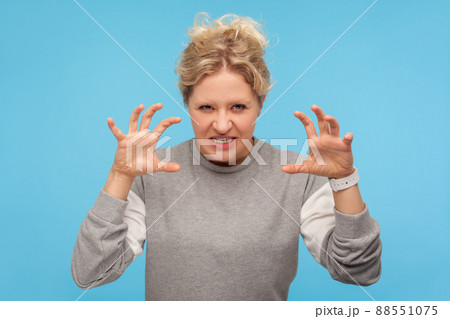 Furious woman with curly hair in sweatshirt standing with raised claws and clenched teeth, experiencing strong anger, irritation, showing aggressive expression. indoor studio shot, blue background 88551075