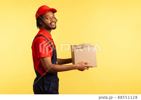 Side view portrait of smiling courier holding cardboard parcel, delivering order door-to-door, shipment and cargo transportation service. Indoor studio shot isolated on yellow background. 88551128