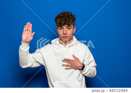 Young handsome man with curly hair wearing casual sweater over blue background Swearing with hand on chest and open palm, making a loyalty promise oath. Young handsome man with curly hair wearing casual sweater over blue background Swearing with hand on chest and open palm, making a loyalty promise oath. 88552994