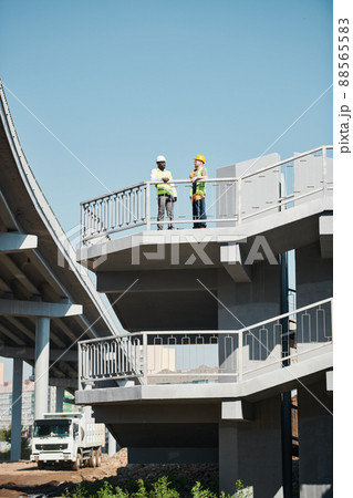 Engineers in white hardhats and safety work vests discussing building project while viewing blueprint at construction site of apartment building Engineers in white hardhats and safety work vests discussing building project while viewing blueprint at construction site of apartment building 88565583