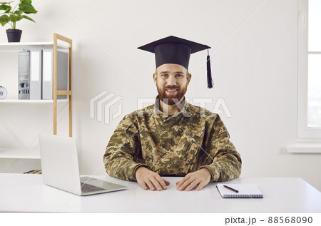 Happy young student in camouflage uniform and graduate hat sitting at desk and smiling 88568090