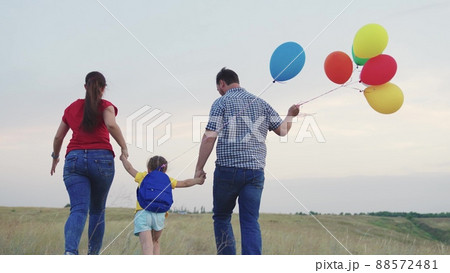 child kid runs with mother father with beautiful colorful balloons. happy family concept. group people running across field with helium colored balloons. happy family running green field together. 88572481