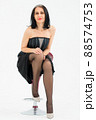 Middle aged woman starting getting grey-haired is posing in studio while sitting on the chair in black dress and stockings on white background, middle age skincare cosmetics, cosmetology concept 88574753