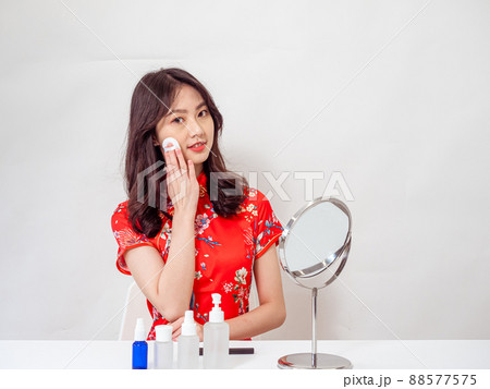Portrait of a young asian woman in traditional cheongsam qipao dress looks at mirror and does skin care isolated on white background. Portrait of a young asian woman in traditional cheongsam qipao dress looks at mirror and does skin care isolated on white background. 88577575
