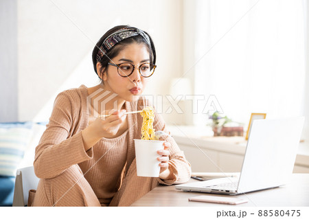 ノートパソコンを見ながらカップラーメンを食べる若い女性 88580475