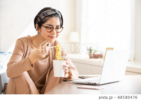ノートパソコンを見ながらカップラーメンを食べる若い女性 88580476