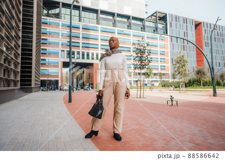 Full body of well dressed African American female in Muslim headscarf standing near modern glass building in downtown and looking away 88586642