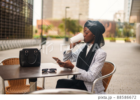 Serious African American female in trendy formal wear and traditional headscarf sitting at table and drinking takeaway coffee while surfing Internet on cellphone 88586672