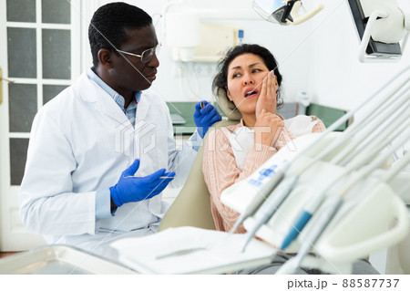 Woman patient who came to an appointment with a male dentist is sitting in a dental chair in clinic office, showing which side of her tooth aches Woman patient who came to an appointment with a male dentist is sitting in a dental chair in clinic office, showing which side of her tooth aches 88587737