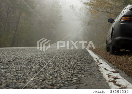 A foggy morning on a country road in an autumn forest. A black car stands on the side of the road. Asphalt road with cracked white markings goes down. A foggy morning on a country road in an autumn forest. A black car stands on the side of the road. Asphalt road with cracked white markings goes down. 88595556