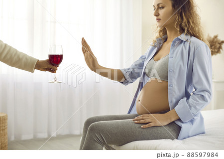 Pregnant woman being offered glass of wine doing stop gesture and refusing to drink 88597984