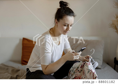 Young woman sitting on the bed at home and taking photo of her old clothes to sell them online. Selling on website, e-commerce. Reuse, second-hand concept. Conscious consumer, sustainable lifestyle. Young woman sitting on the bed at home and taking photo of her old clothes to sell them online. Selling on website, e-commerce. Reuse, second-hand concept. Conscious consumer, sustainable lifestyle. 88601182