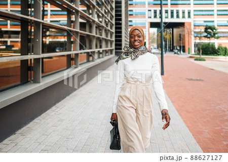 Confident African American female wearing stylish light clothes and hijab walking past modern office towers and looking away Confident African American female wearing stylish light clothes and hijab walking past modern office towers and looking away 88627127