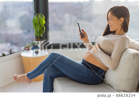 Pregnancy and maternity phone app for online prenatal class, preparation for birth. Happy Asian pregnant woman using cellphone at home watching videos on smartphone 88628254