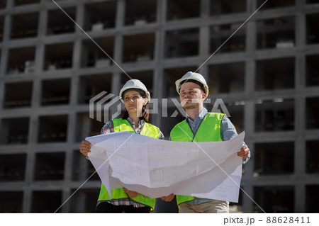 two engineers, a man and a woman in white helmets and protective vests, work at the construction site.  88628411