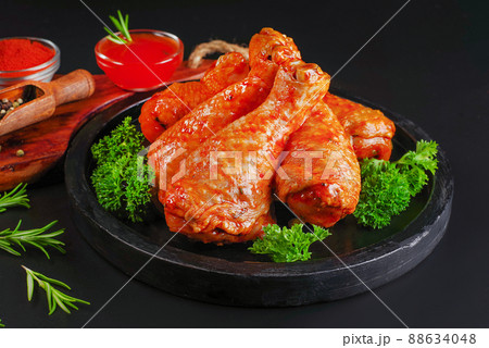 Raw chicken legs marinated in red sauce, on a dark round plate, on a dark background, with herbs, red hot pepper tomatoes and garlic. Chicken legs semi-cooked. Top of view. Raw chicken marinated. 88634048