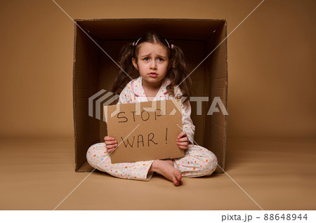 Upset little girl sits inside a cardboard box and shows a Stop the War poster. Social advertisement and concept of a ceasefire, the problems of migrants and refugees during the military conflict 88648944
