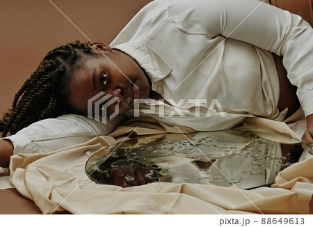 Portrait of African young woman lying near the mirror with water drops on surface and looking at camera Portrait of African young woman lying near the mirror with water drops on surface and looking at camera 88649613
