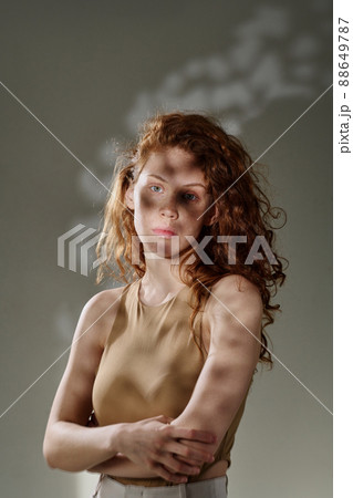 Portrait of young girl with red long curly hair looking at camera standing against the grey background 88649787