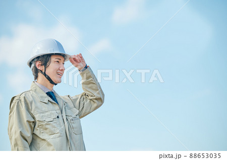 青空を背景にヘルメットをかぶって立つ作業服姿の若い男性 88653035