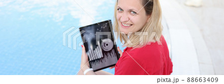 Young woman sitting on side of pool and holding digital tablet with charts in her hands 88663409