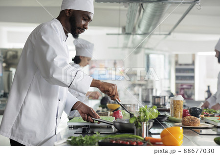 Food industry worker preparing meal in restaurant professional kitchen. Skilled head chef stirring with spatula in pan while cooking gourmet dish for dinner service, garnished with fresh herbs. Food industry worker preparing meal in restaurant professional kitchen. Skilled head chef stirring with spatula in pan while cooking gourmet dish for dinner service, garnished with fresh herbs. 88677030