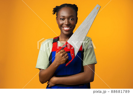 Portrait of a cute african american woman wearing uniform posing with a wood saw in her hands against yellow background 88681789