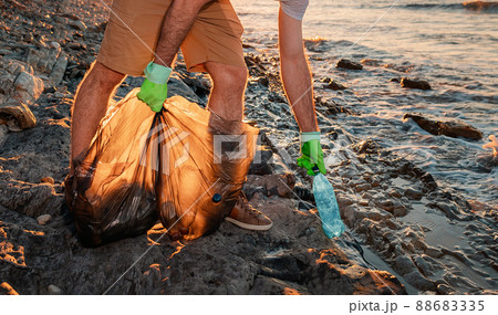 A male volunteer holding plastic bags with garbage and pick ups plastic bottle. Close up. The concept of coastal zone cleaning and world environment day 88683335