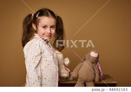 Close-up cute baby girl with two ponytails holding her plush toys and cutely smiles confidently looking at camera, isolated over beige background with copy ad space 88692163