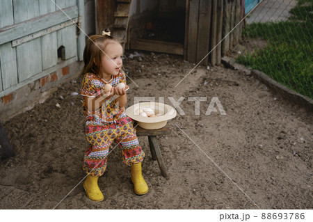Baby is sitting on a bench by the chicken coop and holding fresh eggs in her hands. Next to the girl is a hat filled with chicken eggs from a laying hen. The child looks towards copy space 88693786
