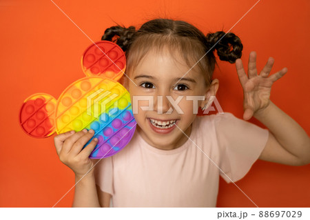 An emotional portrait of a cute little girl on an orange background with a funny hairstyle made of pigtails and a popit toy in her hands. Hand gestures. Pop it toy 88697029