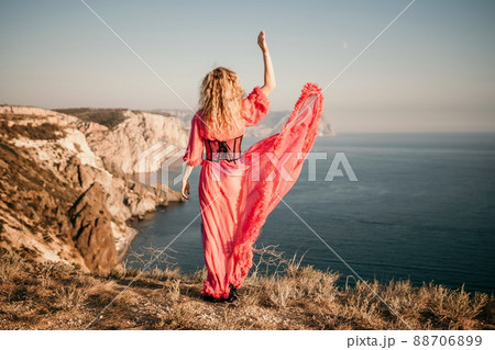 beautiful young caucasian woman with curly blond hair and freckles. Cute woman portrait in a pink long dress posing on a volcanic rock high above the sea beautiful young caucasian woman with curly blond hair and freckles. Cute woman portrait in a pink long dress posing on a volcanic rock high above the sea 88706899