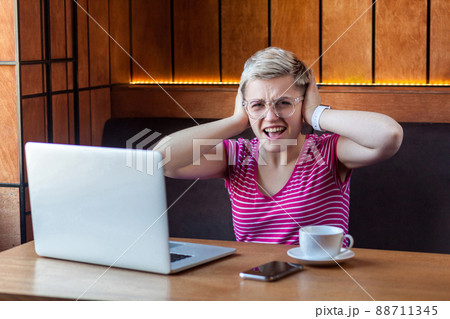 Portrait of attractive young girl freelancer in pink t-shirt is sitting in cafe and working on laptop, covering ears with fingers that not hear to companion. looking at camera 88711345