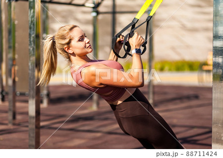 Closeup strong athlete blond woman training outdoor, pulling trx suspension straps, doing upper body workouts for muscle build, fat burning, cardio improvement. Health care and sports activity concept Closeup strong athlete blond woman training outdoor, pulling trx suspension straps, doing upper body workouts for muscle build, fat burning, cardio improvement. Health care and sports activity concept 88711424