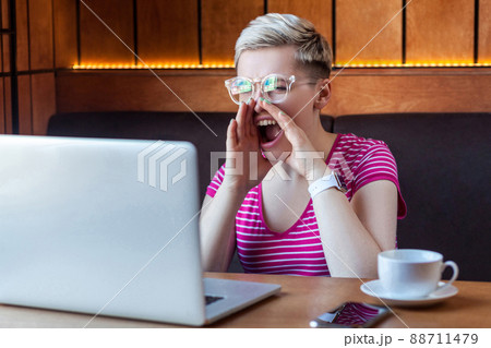 Portrait of angry upset young adult woman with blonde short hair, in pink t-shirt and eyeglasses is sitting in cafe and screaming loud voice to laptop with aggressive face. Indoor, lifestyle Portrait of angry upset young adult woman with blonde short hair, in pink t-shirt and eyeglasses is sitting in cafe and screaming loud voice to laptop with aggressive face. Indoor, lifestyle 88711479