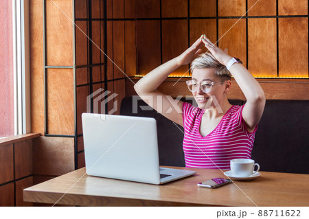 I'm in safety. Portrait of happy young girl with short hair in pink t-shirt and eyeglasses is sitting in office and holding hand upper hand, making roof with hands, looking at laptop, toothy smile I'm in safety. Portrait of happy young girl with short hair in pink t-shirt and eyeglasses is sitting in office and holding hand upper hand, making roof with hands, looking at laptop, toothy smile 88711622