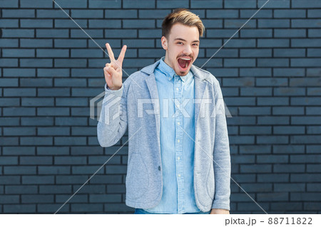 Portrait of surprised handsome young blonde man in casual style standing with victory or peace gesture and looking at camera with amazed happy face. indoor studio shot on brick wall background. Portrait of surprised handsome young blonde man in casual style standing with victory or peace gesture and looking at camera with amazed happy face. indoor studio shot on brick wall background. 88711822